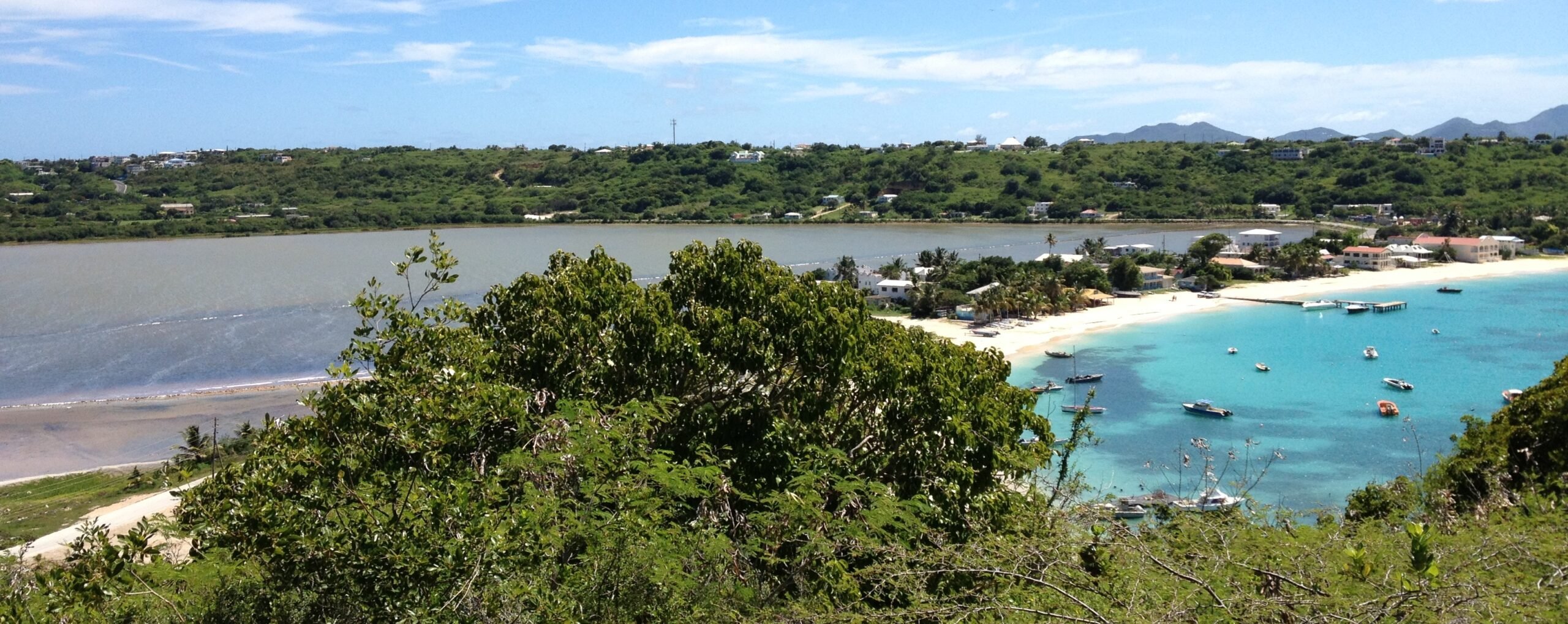 Elevated view overlooking Salt Road Pond and coastline in Anguilla, showing turquoise waters, sandy beach, coastal homes, and surrounding green hills under a bright blue sky.
